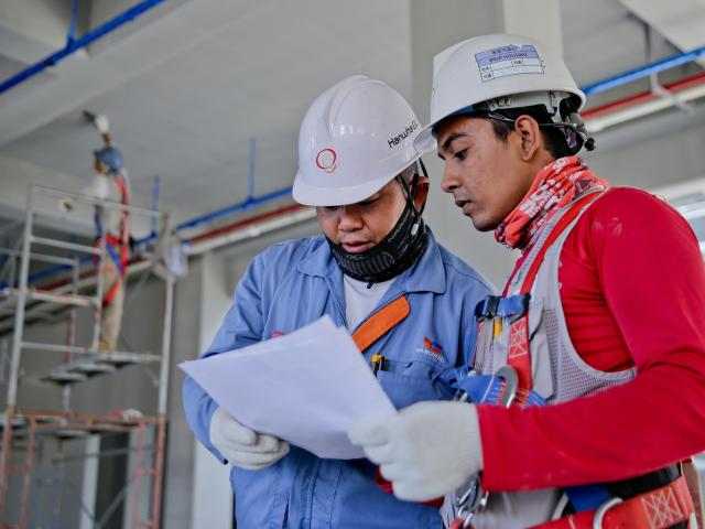 Two men in hard hats look at a piece of paper. In the background, construction is underway.